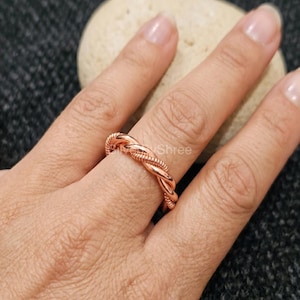 Könnte beinhalten: Ein kupferfarbener Ring mit geflochtenem Design. Der Ring wird an einem Finger getragen, wobei das Wort "Shree" auf dem Ring sichtbar ist. Der Ring besteht aus glänzendem Metall und hat ein gedrehtes Seil-Aussehen.