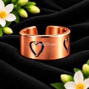 May include: A copper-toned open band ring featuring a brushed metallic finish and engraved heart outlines. The text SilverByShree is visible on the front surface of the ring. The band has a wide, flat profile with a gap at the back for adjustment.