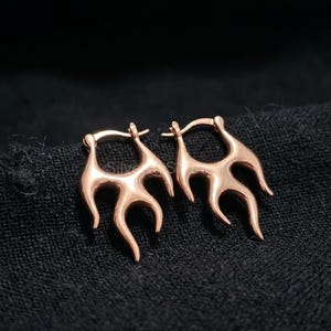 Puede incluir: Un par de pendientes de aro en forma de llama de color oro rosa. Los pendientes tienen un acabado pulido y un diseño único, que se asemeja a llamas estilizadas. Se muestran sobre un fondo de tela oscura.