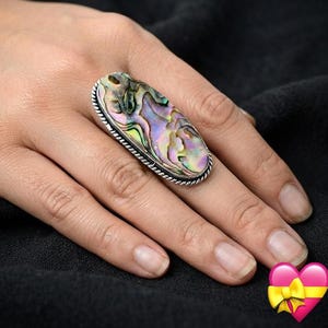Può includere: Un anello di forma ovale con un grande intarsio in conchiglia di abalone iridescente con motivi vorticosi nei toni del verde, viola e rosa. La conchiglia è incastonata in una montatura in metallo color argento lucido con una trama a corda.