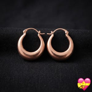 May include: A pair of rose gold hoop earrings with a thick, rounded profile and a polished finish. The earrings feature a hinged post mechanism at the top, designed for secure closure. The metal has a warm, pinkish-gold hue and a smooth surface.