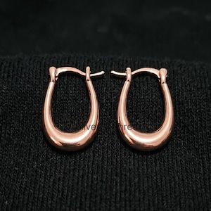 May include: A pair of rose gold hoop earrings. The earrings have a smooth, polished finish and a classic oval shape. The earrings are displayed on a dark background.