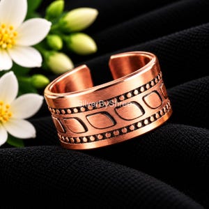 May include: A wide copper ring featuring an open band design with a brushed finish. The surface is decorated with a central row of repeating leaf-like shapes bordered by two rows of small, raised dots. The text SilverByShree is visible.