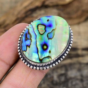 Può includere: Anello ovale con conchiglia di abalone e fascia argentata. Il guscio iridescente presenta sfumature di blu, verde e bianco. La fascia dell'anello ha un design a perline attorno al guscio. Gioielli.