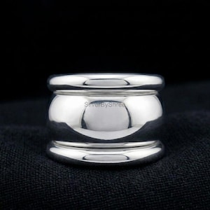 Può includere: Un anello in argento lucido con una fascia larga. L'anello ha una parte superiore e inferiore arrotondata con una sezione centrale più ampia e curva. Viene mostrato su una superficie scura e strutturata. Il testo "SilverByShree" è visibile.