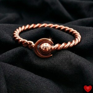 Puede incluir: Un anillo de metal trenzado en tono cobre con un elemento circular central enmarcado por una forma de media luna. La banda tiene una textura continua similar a una cuerda y el punto focal consiste en una esfera lisa dentro de un engaste curvo.