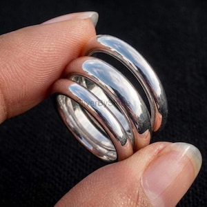 Può includere: Un set di tre anelli in argento lucido con fasce lisce e arrotondate. Gli anelli sono impilati insieme, mostrando una superficie metallica riflettente e un design circolare semplice e continuo. Il metallo presenta una finitura brillante.