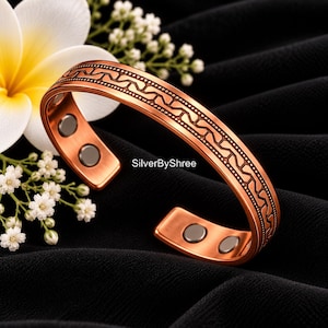 May include: A copper-colored cuff bracelet featuring a repeating geometric pattern along the outer band and four circular magnets embedded on the inner surface. The text SilverByShree is visible on the metal surface of the open-ended bracelet.
