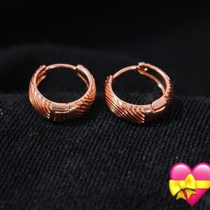 Könnte beinhalten: Ein Paar Creolen aus Roségold mit einer strukturierten Oberfläche und diagonalen Rillen. Die kleinen, kreisförmigen Ohrringe verfügen über einen Klappverschluss und eine polierte Metalloberfläche, die das Licht reflektiert.