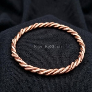 May include: A copper-colored twisted wire bracelet. The bracelet is circular and appears to be made of multiple strands of wire woven together. The background is a dark fabric. The text "SilverByShree" is visible.