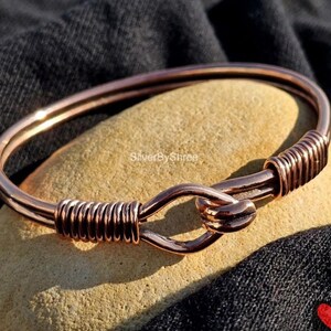 May include: A copper-colored wire bangle bracelet featuring a hook-and-eye style closure mechanism. The design includes two sections of tightly coiled wire wrapping around the main band on either side of the central clasp. The text SilverByShree is visible.