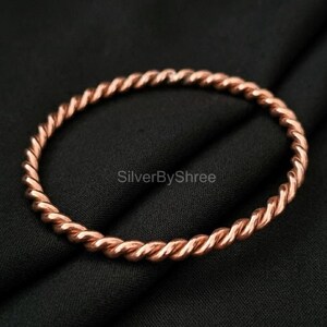 May include: A copper-colored twisted bangle bracelet. The bracelet has a rope-like design and is displayed on a black background. The jewelry is a simple, elegant accessory.