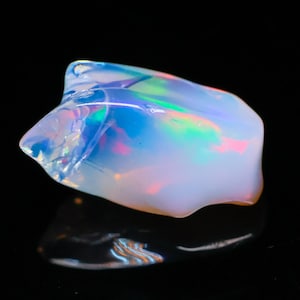 May include: A polished opal gemstone with a milky white base color and iridescent flashes of blue, green, and pink. The stone has a smooth, rounded shape with a reflective surface, set against a black background.