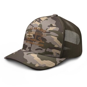 May include: A camouflage trucker hat with a brown mesh back and a curved brim. The front of the hat features the text "U.S. AIR FORCE" above a graphic of a loader, with the text "60K TUNNER LOADER" below.