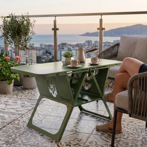 Op de afbeelding: Een groene metalen klaptafel met een hertenkopontwerp en het woord "Fiways" staat op een balkon. Twee kopjes en een koffiepot staan op de tafel. Een persoon zit in een rieten stoel.