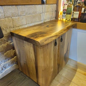 May include: A wooden cabinet with a live edge top and two doors. The cabinet is made of light-coloured wood with a natural grain pattern. The doors have black metal handles. The cabinet is next to a bar.