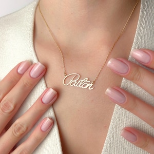 Può includere: Una collana color oro con il nome "Paiton" in corsivo. La collana è indossata intorno al collo. L'immagine mostra la collana e la parte superiore del petto.