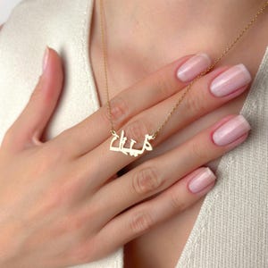 May include: A gold-colored necklace with an Arabic name pendant. The pendant is in a stylized script and hangs from a delicate chain. The necklace is displayed on a person's chest, with their hand gently touching the chain.