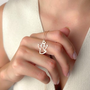 Könnte beinhalten: Eine goldfarbene Engels-Anhänger-Halskette. Der Anhänger hat ein ausgeschnittenes Design eines Engels mit Flügeln und einem Heiligenschein. Die Halskette wird auf der Hand einer Person präsentiert und zeigt die zarte Kette und den Anhänger.