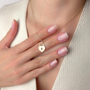 Op de afbeelding: Een goudkleurige ketting met een hartvormige hangerslot hanger. De hanger heeft een sleutelgat detail. De ketting wordt om de nek van een persoon gedragen en de hand raakt de hanger aan. De achtergrond is een lichtgekleurde top.