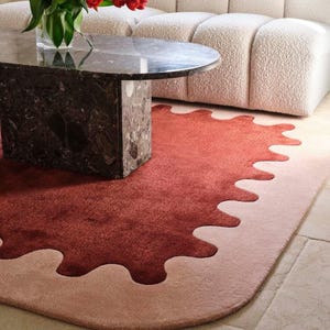 May include: A modern living room scene featuring a round marble coffee table and a plush, cream-colored sofa. A vibrant red and pink rug with a unique, organic shape sits on the floor, adding a pop of color. A vase of red flowers adds a touch of nature.