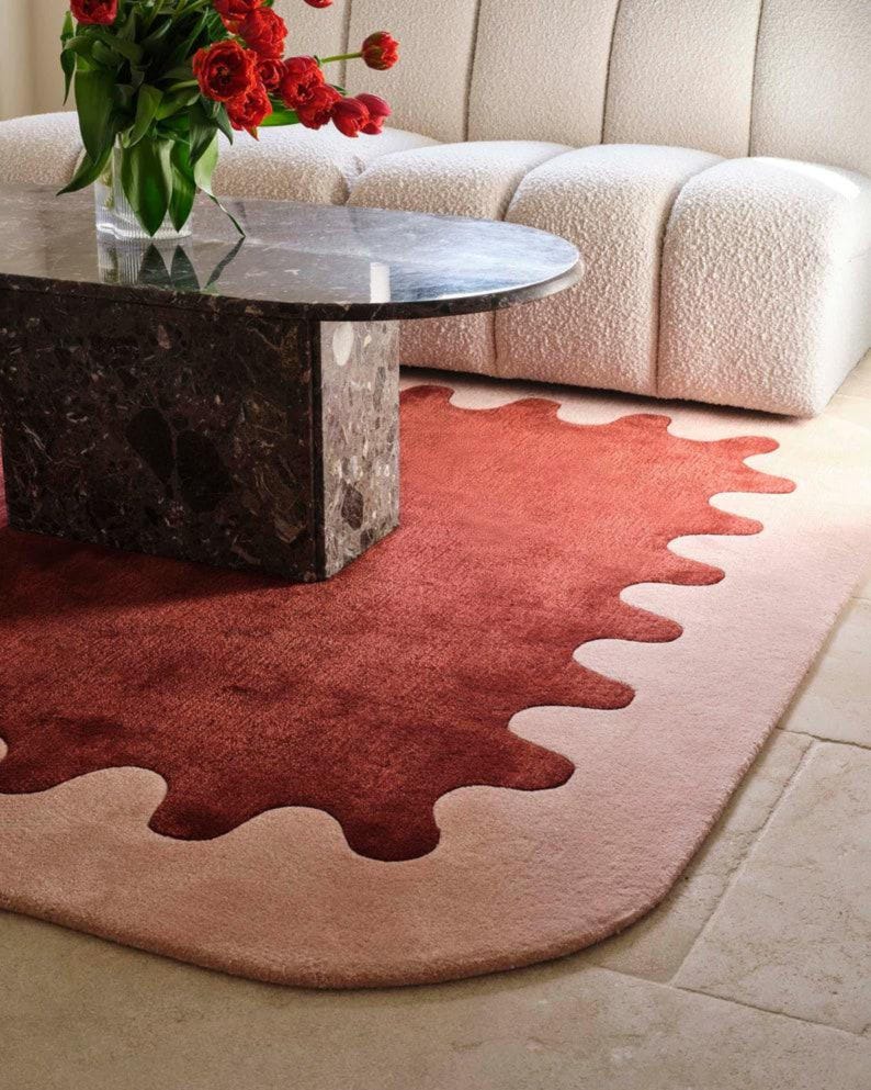 May include: A modern living room scene featuring a round marble coffee table and a plush, cream-colored sofa. A vibrant red and pink rug with a unique, organic shape sits on the floor, adding a pop of color. A vase of red flowers adds a touch of nature.
