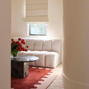 May include: A living room scene featuring a cream-colored, textured sofa, a round marble coffee table, and a red and pink rug with a unique wavy edge. A vase of red flowers sits on the table. A window with a beige shade is visible.