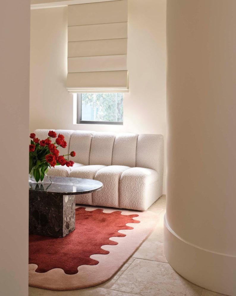 May include: A living room scene featuring a cream-colored, textured sofa, a round marble coffee table, and a red and pink rug with a unique wavy edge. A vase of red flowers sits on the table. A window with a beige shade is visible.
