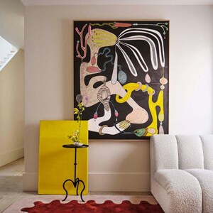 May include: An abstract painting with black background and colorful shapes hangs above a white sofa. A yellow canvas and a small black table with a plant sit next to a red rug with an irregular edge. The room has a modern, artistic aesthetic.