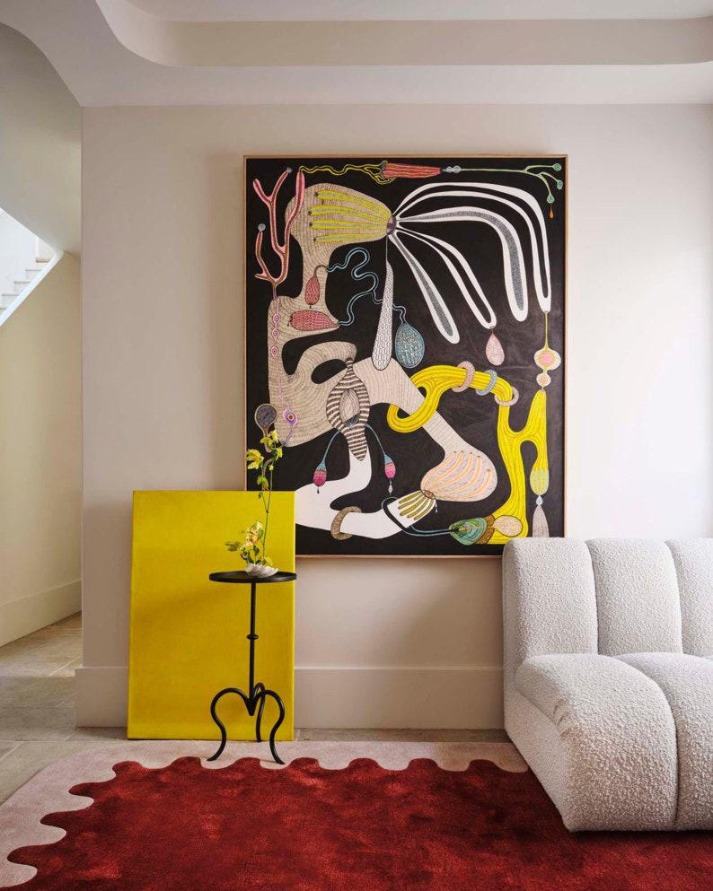 May include: An abstract painting with black background and colorful shapes hangs above a white sofa. A yellow canvas and a small black table with a plant sit next to a red rug with an irregular edge. The room has a modern, artistic aesthetic.