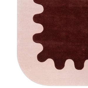 May include: A modern rug with a unique design. The rug features a light pink border with a dark brown, scalloped shape in the center. The rug's corner is rounded, adding to its contemporary aesthetic.