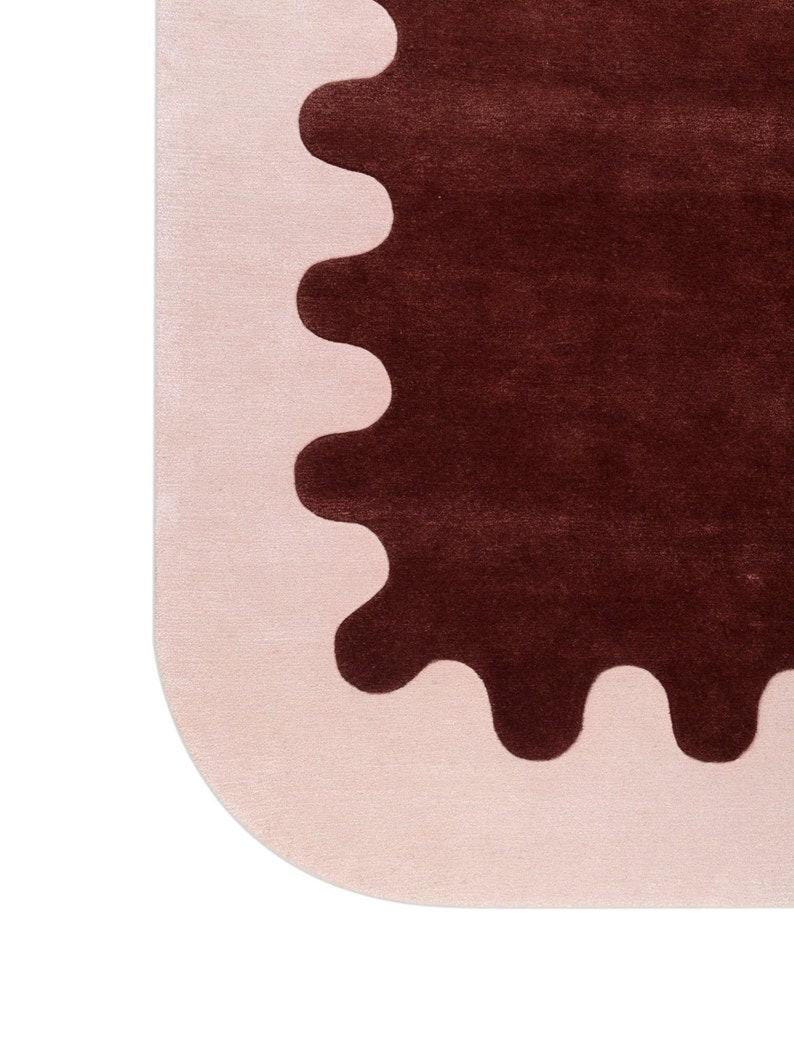May include: A modern rug with a unique design. The rug features a light pink border with a dark brown, scalloped shape in the center. The rug's corner is rounded, adding to its contemporary aesthetic.