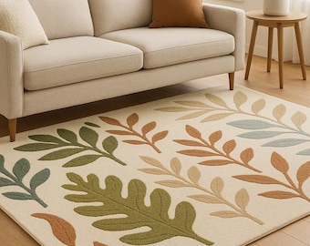 May include: A cream-colored area rug with a leaf pattern in shades of green, brown, and blue. The rug is placed under a beige sofa with brown and cream-colored pillows. A wooden side table and lamp are visible.