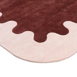 May include: A close-up of a rug with a unique design. The rug features a light pink border with a dark brown, irregular shape in the center. The rug has a soft, plush texture and a modern aesthetic.