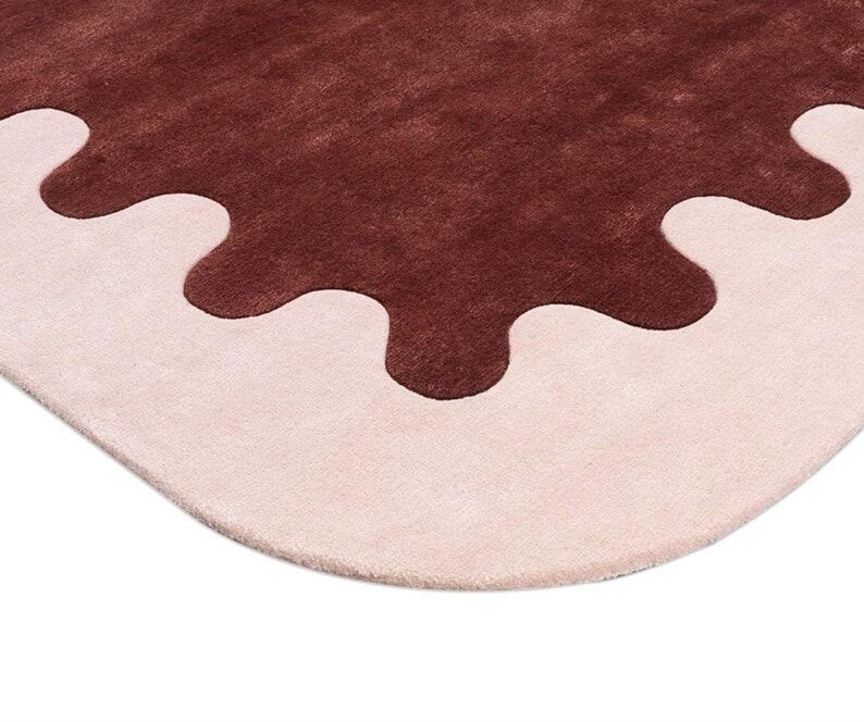 May include: A close-up of a rug with a unique design. The rug features a light pink border with a dark brown, irregular shape in the center. The rug has a soft, plush texture and a modern aesthetic.