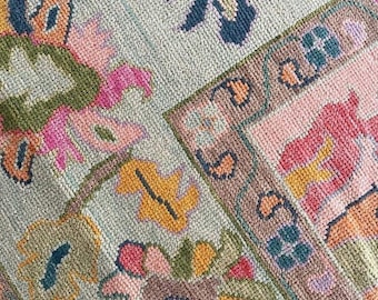 Può includere: Primo piano di una sezione di un tappeto con sfondo verde chiaro e motivo floreale ripetuto. Il tappeto presenta disegni floreali rosa, gialli, blu e verdi, con un bordo marrone. Il tappeto ha un aspetto strutturato.