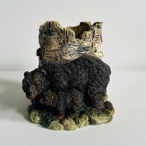 May include: A detailed figurine of a bear and cub, with a textured, dark brown fur. The bear is positioned in front of a tree stump-shaped container. The base is a mix of green and tan, suggesting a forest setting. The figurine is made of a resin-like material.