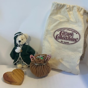 Może przedstawiać: Kolekcja przedmiotów Cottage Collectibles, w tym pluszowy miś w zielonym płaszczu, drewniane serce, mała brązowa torba z cukrowymi laskami i płócienna torba z napisem "Artist Designed Cottage Collectibles by Ganz."