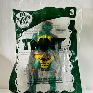 May include: A green Raphael action figure from the Teenage Mutant Ninja Turtles franchise, packaged in a clear plastic bag. The bag has the Happy Meal logo and the number 3. The figure is wearing a red mask and yellow belt.