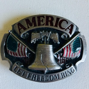 May include: An oval metal belt buckle featuring a central Liberty Bell icon flanked by two crossed American flags. The word AMERICA is embossed at the top, and the phrase LET FREEDOM RING is embossed at the bottom in silver-toned metal.