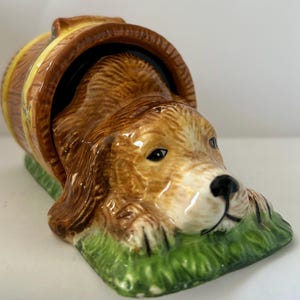 Può includere: Statuetta di cane in ceramica che esce da una botte di legno. Il cane ha pelo marrone, occhi neri e naso nero. La botte è marrone con dettagli gialli. La base è verde, simile all'erba.