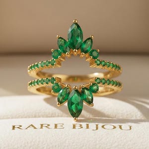Peut inclure: Une bague en or ornée de pierres précieuses vert émeraude en forme de couronne, présentée dans un écrin à bijoux. La bague a un double anneau avec de petites pierres vertes. La boîte est marron avec l'inscription "RARE BIJOU".