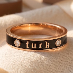 May include: A rose gold band ring featuring a black enamel inlay with the word "fuck" written in a gothic font. The ring is accented with two small, round, clear stones set on either side of the text, creating a distinct contrast in color.