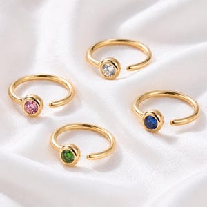 May include: Four gold-colored rings with open bands and bezel-set gemstones. The gemstones are pink, clear, green, and blue. The rings are displayed on a white, silky surface.