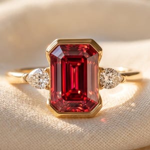 Könnte beinhalten: Ein Goldring mit einem großen, rechteckigen, smaragdgeschliffenen roten Edelstein in der Mitte, flankiert von zwei kleineren, birnenförmigen klaren Edelsteinen auf beiden Seiten. Der Ring ist schmal und poliert und hält die Steine in einer Zargenfassung.