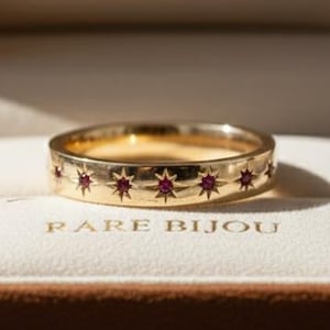Op de afbeelding: Een gouden ring met een sterrenpatroon en kleine rode edelstenen, gepresenteerd in een open bruine fluwelen sieradendoos. De ring ligt op een witte ondergrond. De tekst "RARE BIJOU" is zichtbaar.