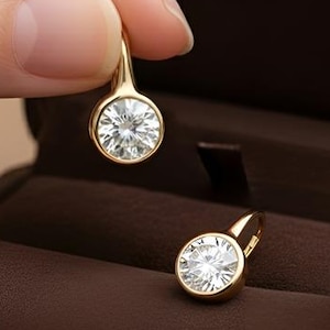 K&ouml;nnte beinhalten: Ein Paar goldfarbene Ohrringe mit runden, klaren Edelsteinen. Ein Ohrring wird von einer Hand gehalten, der andere liegt in einer braunen Schmuckschatulle. Die Schatulle ist ge&ouml;ffnet und zeigt die Ohrringe. Der Text "RARE BIJOU" ist sichtbar.