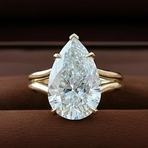 Puede incluir: Un anillo de oro con un diamante en forma de pera, presentado en una caja de joyería de terciopelo marrón. La caja está abierta, revelando el anillo contra un interior marrón oscuro. El texto "RARE BIJOU" es visible en la caja. Una joya de lujo.