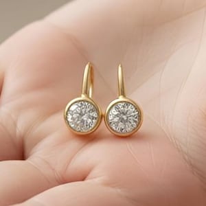 Op de afbeelding: Een paar goudkleurige oorbellen met een ronde, heldere edelsteen in een bezel. De oorbellen worden in een open hand getoond, waarbij hun ontwerp wordt benadrukt. Een klassiek sieraad.