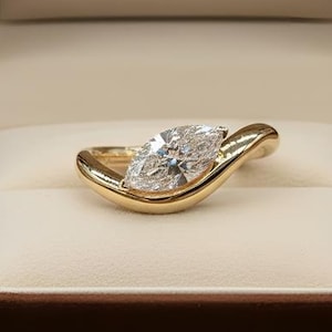 Può includere: Anello in oro con diamante taglio marquise, presentato in un cofanetto di velluto marrone aperto. L'interno del cofanetto è color crema e sul coperchio è stampata la scritta "RARE BIJOU".